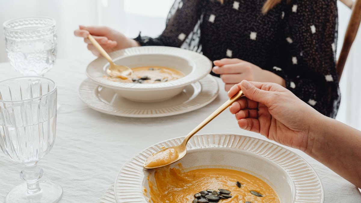 A close-up of hands enjoying creamy pumpkin soup at an elegant dining table setting.
